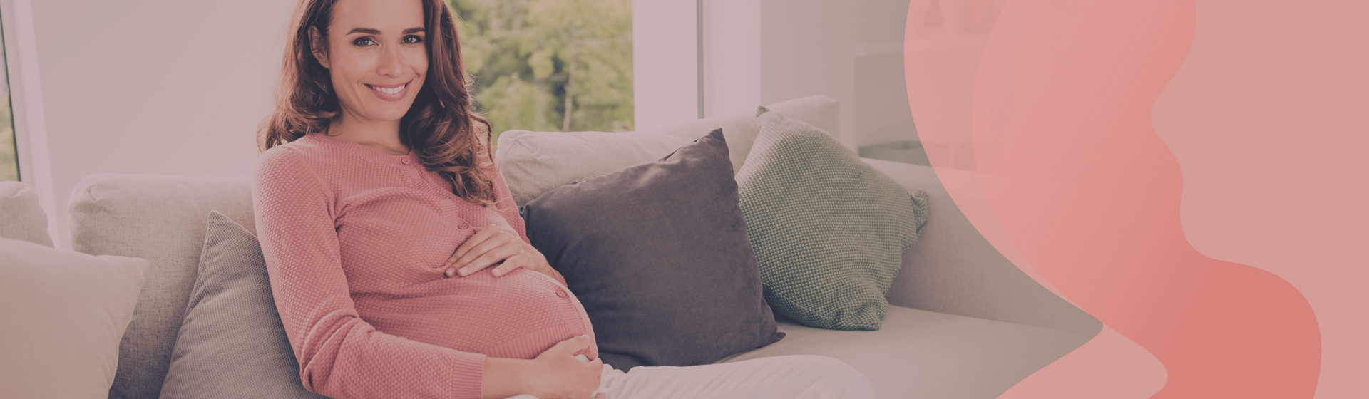 Pregnat woman sitting on a couch, holding her belly and smiling at the camera.